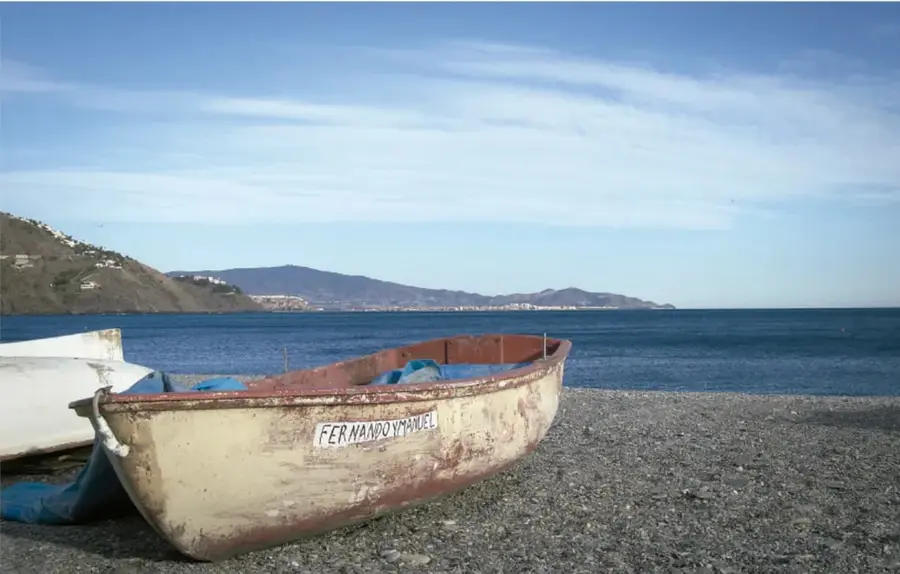 Fotografía de una playa con un bote de madera viejo en la orilla, el mar azul y montañas al fondo; el bote tiene escrito 'FERNANDO Y MANUEL'.