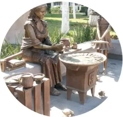 Fotografía de un monumento de bronce en una plaza: una mujer sentada hace tortillas sobre un comal, acompañada de vasijas y una niña, en homenaje a las enchiladas potosinas.