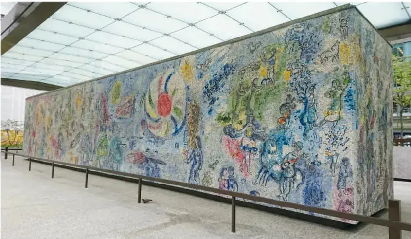 Fotografía de un gran mural de Marc Chagall, de forma alargada y estructura cúbica, cubierto por una composición colorida con figuras humanas, animales y símbolos sobre fondo azul. Está instalado bajo una cubierta de vidrio y rodeado por una baranda metálica en un espacio público o museístico.