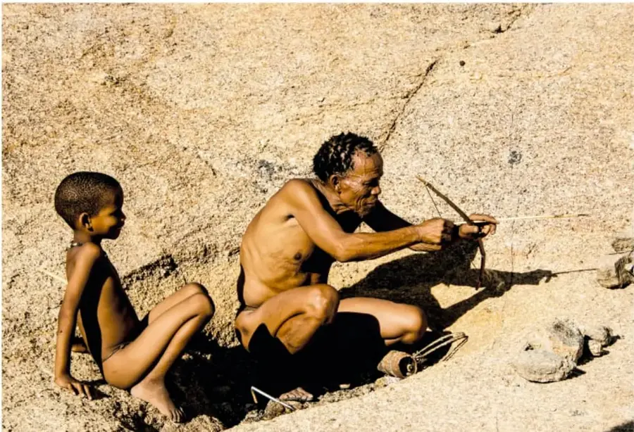 Fotografía de un hombre adulto y un niño sentados sobre arena, mientras el adulto manipula un arco y flecha; evoca la vida de caza y recolección de la prehistoria.