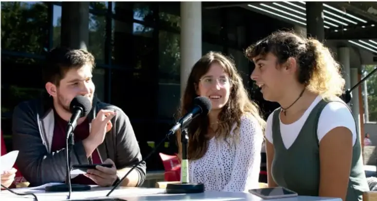 Fotografía de tres estudiantes sentados frente a micrófonos, aparentemente en una transmisión o rueda de prensa estudiantil, dando a conocer los resultados de su proyecto.