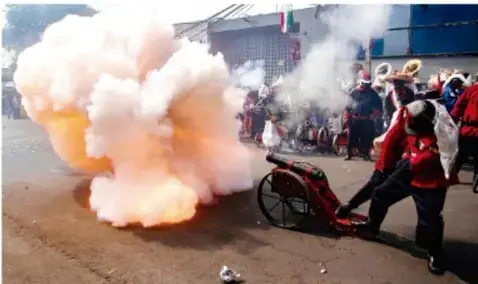 Foto de una calle con humo y música tradicional, posiblemente conmemoración cívica o desfile.