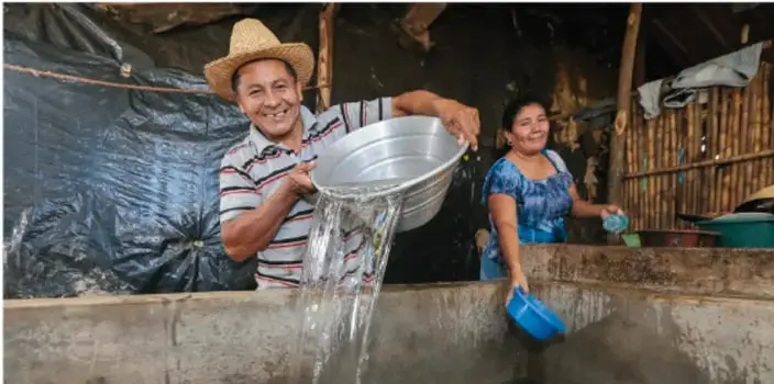 Dos personas, un hombre con sombrero y una mujer, vierten agua de un balde metálico en un contenedor de concreto; escena rural relacionada con el cuidado del agua.