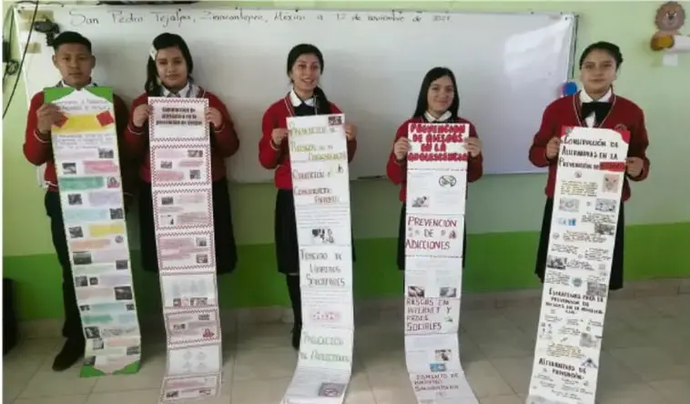 Cinco alumnos de Telesecundaria en uniforme muestran guías de prevención de riesgos largas y plegadas, con títulos como 'Prevención de accidentes', 'Prevención de adicciones' y 'Autoestima'. Pizarrón y cartel institucional al fondo.