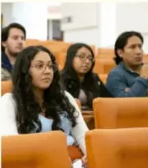 Fotografía de estudiantes sentados en fila en un aula, escuchando una presentación.