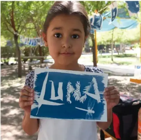 Fotografía de una niña sonriente sosteniendo una pintura azul con letras blancas.