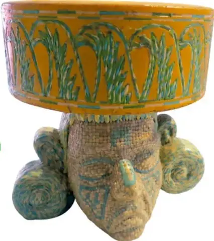 Escultura *Huehuetéotl de sabores II* (2016): figura de un rostro anciano de piedra con un recipiente o brasero de color naranja con motivos verdes sobre la cabeza, rodeado de rollos de colores.