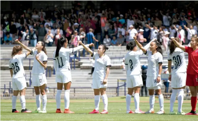 Fotografía de un equipo de fútbol femenino con uniforme blanco en el campo, celebrando y dándose las manos frente a las gradas con público, mientras una jugadora rival con uniforme rojo está al lado.