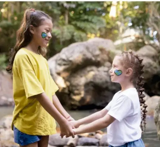 Fotografía de dos niñas tomadas de las manos mirándose sonrientes; una viste de amarillo y la otra tiene pintura verde en la mejilla; simboliza la reconciliación y el acuerdo pacífico.