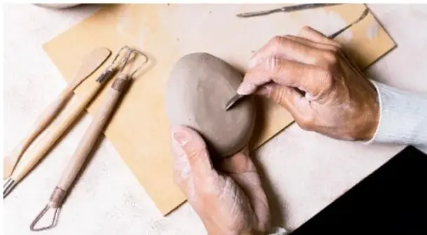 Manos de un artesano trabajando una pieza de barro con herramientas de modelado sobre una mesa de trabajo; se observan espátulas y utensilios de cerámica.