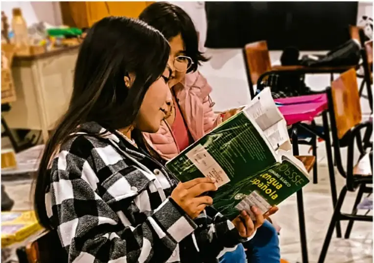 Fotografía de dos estudiantes de telesecundaria leyendo juntas el libro de texto de Lengua Española en el aula; sillas, pupitres y objetos escolares al fondo. Crédito: Luis Eduardo García Meojo, FOTOTECA.