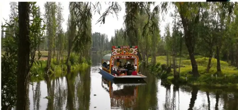 Fotografía de una trajinera con personas navegando por un canal rodeado de árboles y chinampas, paisaje rural de Xochimilco.