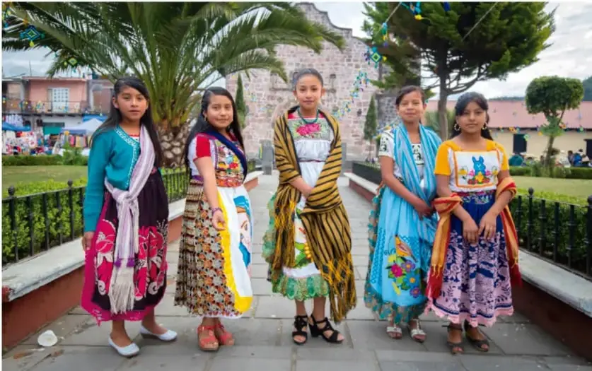 Fotografía de cinco niñas con trajes típicos coloridos de pie en un parque o plaza, posando para la foto; probablemente vinculada a la presentación del proyecto.