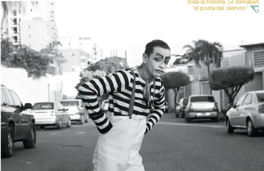Fotografía en blanco y negro de un mimo con camisa a rayas, tirantes y pantalones blancos, en una postura inclinada sobre una calle con autos estacionados y edificios al fondo.