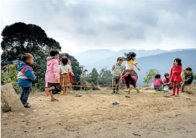Fotografía de niñas y niños jugando al salteador/brinco con un listón en un patio al aire libre con montañas al fondo.