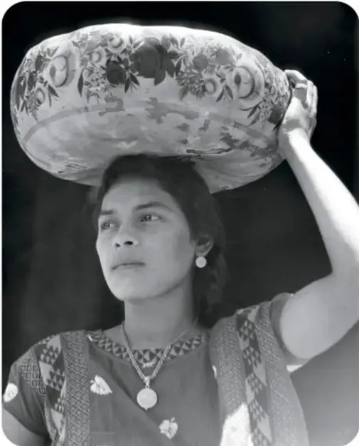 Retrato en blanco y negro de una mujer con una vasija ancha decorada con flores sobre la cabeza, ropa tradicional bordada, aretes, collar y trenza; pie: 'Tehuana con jicara, ca. 1929'.