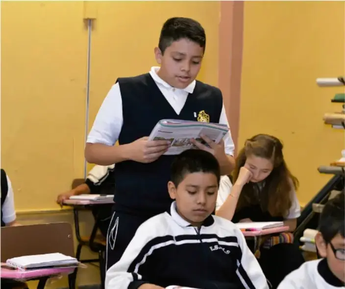 Fotografía de estudiantes en un aula; un estudiante de pie lee en voz alta un libro o cuaderno frente a sus compañeros, ilustrando la exposición en asamblea.
