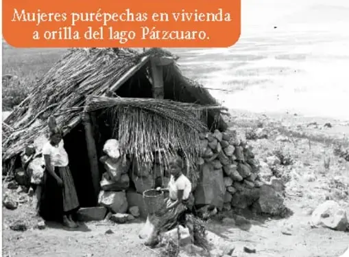 Fotografía en blanco y negro titulada 'Mujeres purépechas en vivienda a orilla del lago Pátzcuaro', donde se observa una casa de adobe con techo de paja y varias mujeres con vestimenta tradicional realizando actividades cotidianas.