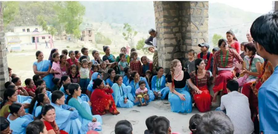 Grupo grande de mujeres y niñas, muchas con ropa azul y roja, sentadas en círculo en un patio de tierra junto a una estructura de piedra, en una reunión comunitaria.