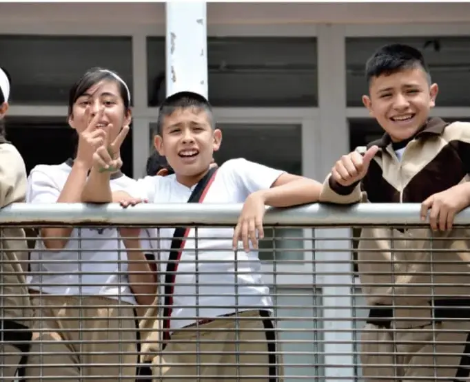 Grupo de estudiantes adolescentes en uniforme blanco y beige asomados a una barandal metálico, sonriendo y haciendo gestos de paz y pulgares arriba.