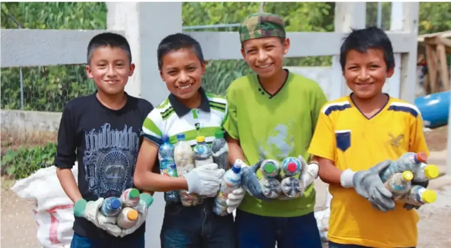 Cuatro estudiantes de primaria sostienen botellas PET rellenas de residuos plásticos (ecoladrillos) y usan guantes; al fondo hay costales con más botellas.