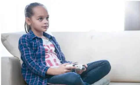 Fotografía de una niña sentada en un sofá sosteniendo un control de videojuego, ilustrando el uso de dispositivos electrónicos para jugar.