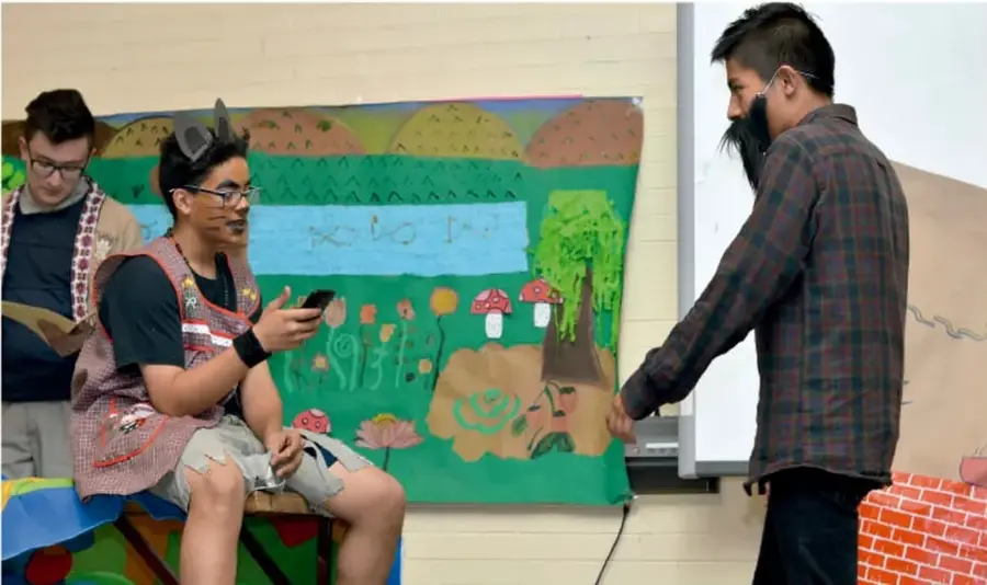 Fotografía de estudiantes presentando un proyecto con un mural pintado de fondo; uno de ellos lee de un teléfono mientras otro explica junto a un pizarrón.