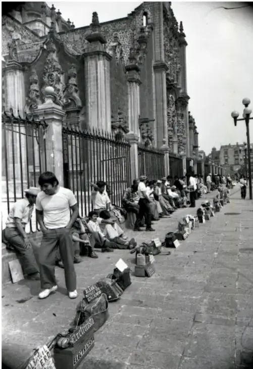Fotografía en blanco y negro de personas sentadas en el suelo a lo largo de una calle, junto a una reja, con mercancía colocada en el piso; parecen vendedores ambulantes.