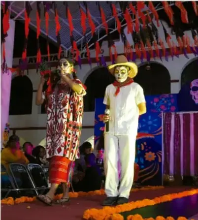 Fotografía de una presentación comunitaria con personas con vestimenta y calaveras tradicionales de día de muertos sobre un escenario con flores de cempasúchil.