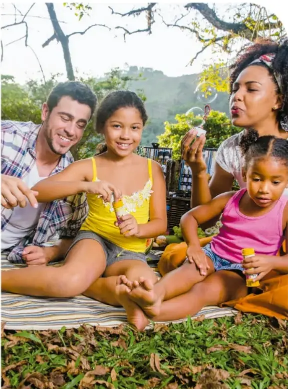 Fotografía de una familia (padre, madre y dos niñas) en un picnic al aire libre, soplando burbujas de jabón, en un entorno natural.