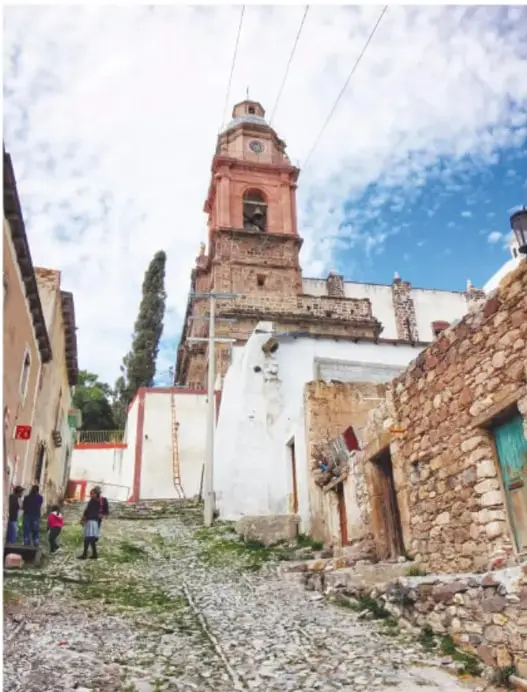 Vista de una calle empedrada hacia la parroquia con su torre rosa/terracota; casas de piedra y adobe, un ciprés alto y visitantes caminando.
