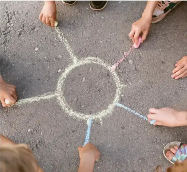 Niños y niñas dibujan con gises de colores un círculo y líneas en el patio, formando una figura similar a una flor o rueda.
