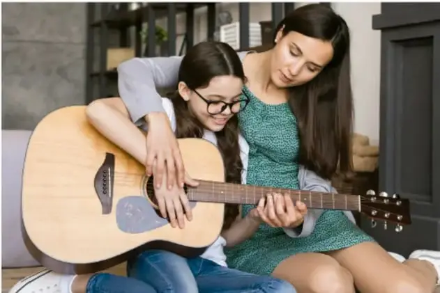 Fotografía de una joven y una niña sentadas en el piso tocando una guitarra acústica, ilustrando la creación musical colectiva.