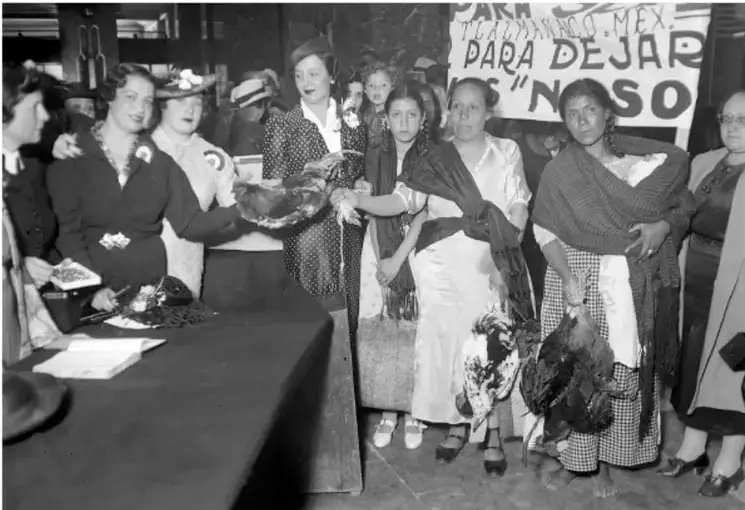 Fotografía en blanco y negro de un grupo de mujeres frente a un puesto de recaudación; sostienen aves y un cartel con la leyenda "PARA DEJAR... NOSO". Representa la participación ciudadana en la colecta para pagar la indemnización de la expropiación petrolera.
