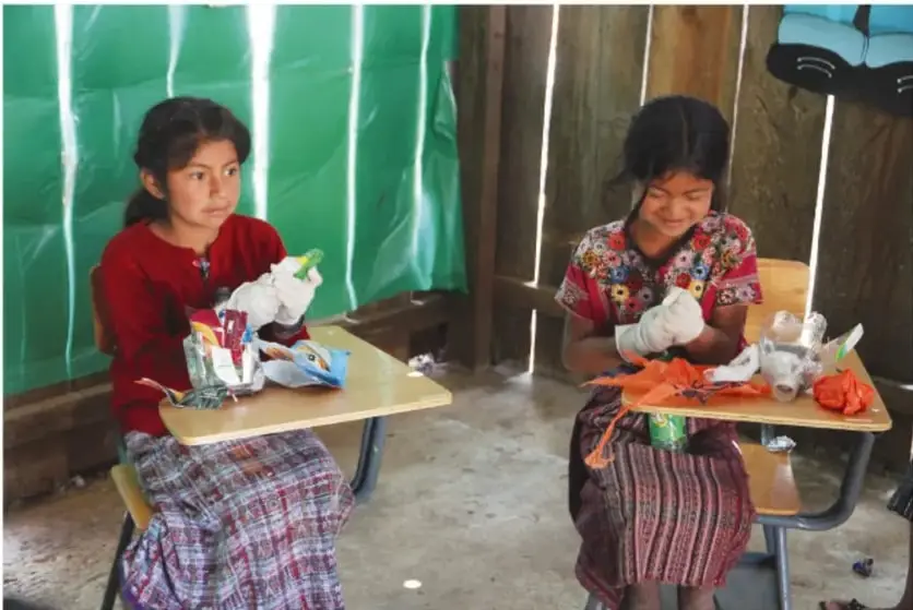 Dos niñas sentadas en sus pupitres forrando botellas para hacer ecoladrillos en un aula con paredes de madera y plástico verde.