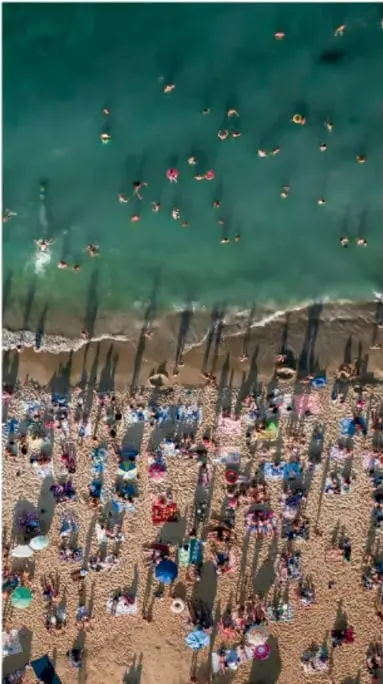Fotografía aérea de una playa muy concurrida con bañistas en el mar y sombrillas en la arena, que ilustra la alta concentración humana.