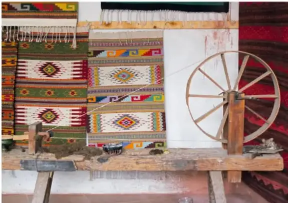Telar de pedales de madera con telas tejidas de motivos geométricos de colores y una rueca.