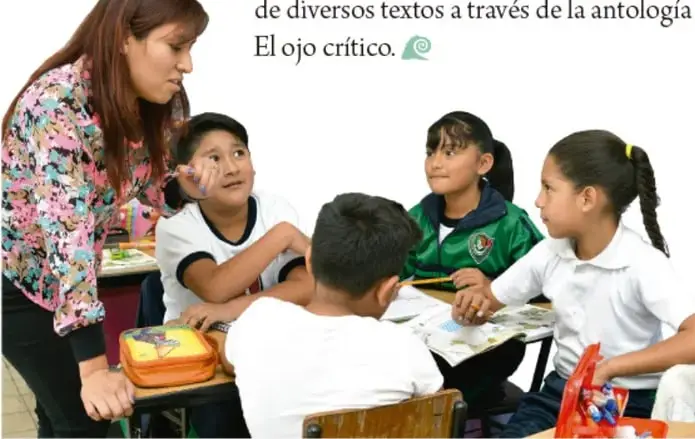 Fotografía de una maestra de pie junto a cuatro estudiantes de primaria sentados en sus pupitres, dialogando en el aula durante una actividad grupal.