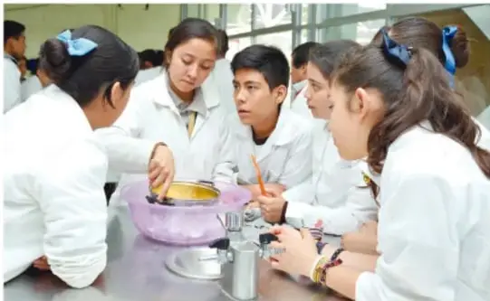 Fotografía de un grupo de estudiantes de secundaria con bata blanca alrededor de una mesa de laboratorio; observan y manipulan material (vasos, recipientes) mientras realizan una actividad experimental.