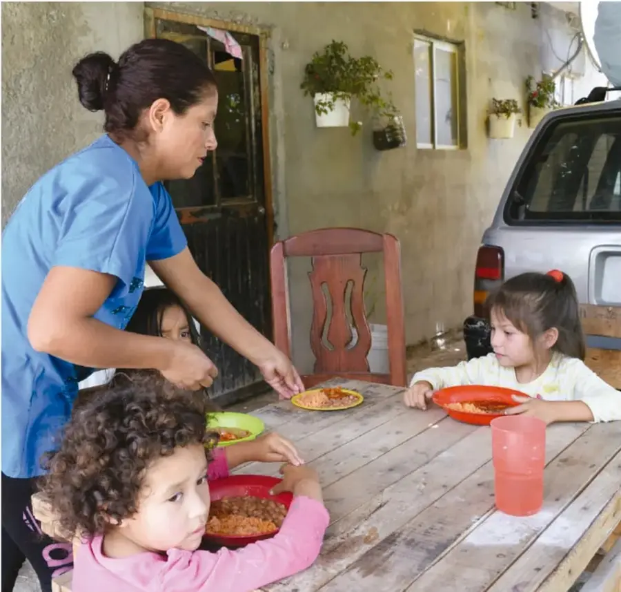 Fotografía de una mujer con uniforme azul que sirve comida a tres niñas sentadas en una mesa de madera, en un patio con una camioneta al fondo.