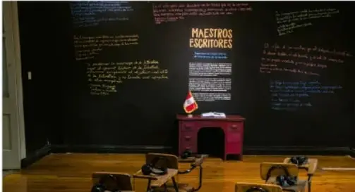Fotografía de un salón de clases con un pizarrón negro con la leyenda "MAESTROS ESCRITORES" y escritos manuscritos, con pupitres de madera en primer plano.