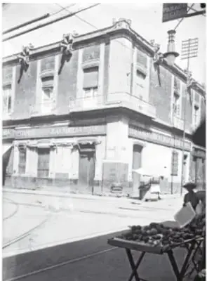 Fotografía histórica en blanco y negro de la misma esquina del edificio en el pasado, con la cabeza de serpiente en la pared y un puesto con canastas en la calle.