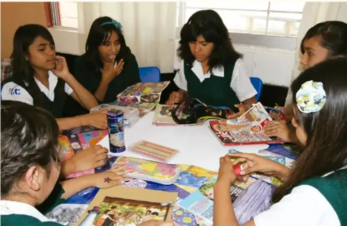 Fotografía de varias estudiantes de secundaria con uniforme, sentadas alrededor de una mesa cubierta de álbumes, revistas y materiales, revisando y comentando sus trabajos.