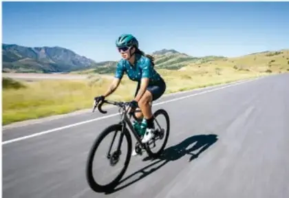 Fotografía de un ciclista con casco y ropa deportiva pedaleando en una carretera rodeada de montañas, bajo un cielo despejado.