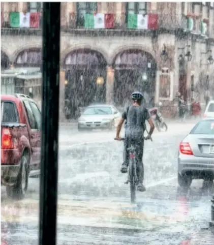 Persona en bicicleta bajo lluvia intensa en una calle con autos y edificios con banderas de México.