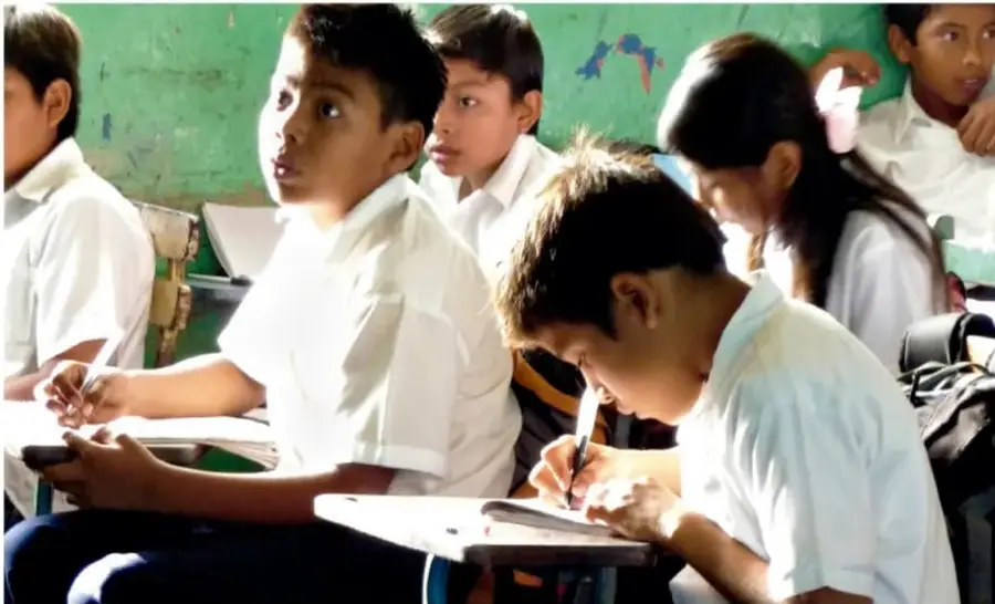 Fotografía de estudiantes de primaria sentados en sus pupitres escribiendo en el cuaderno dentro del salón de clases.