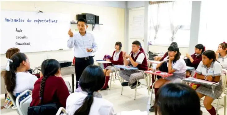Foto de un aula de telesecundaria donde el docente explica frente a una cartelera que dice 'Horizontes de expectativas: Aula, Escuela, Comunidad' y los estudiantes escuchan.