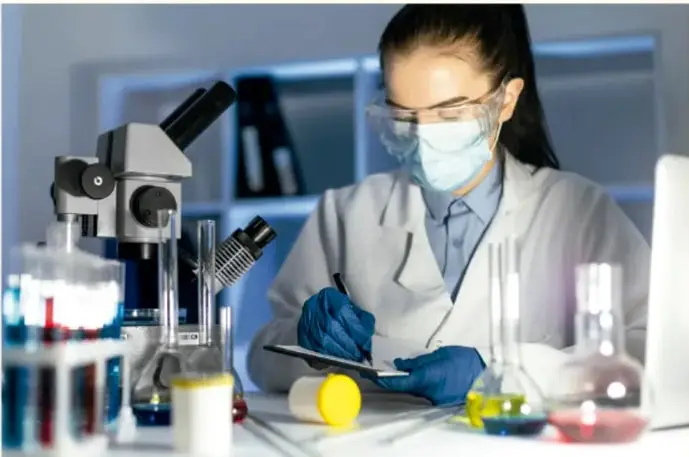 Fotografía de una estudiante con bata blanca, guantes azules, mascarilla y goggles de laboratorio, escribiendo en una tableta frente a un microscopio y material de vidrio con líquidos de colores.
