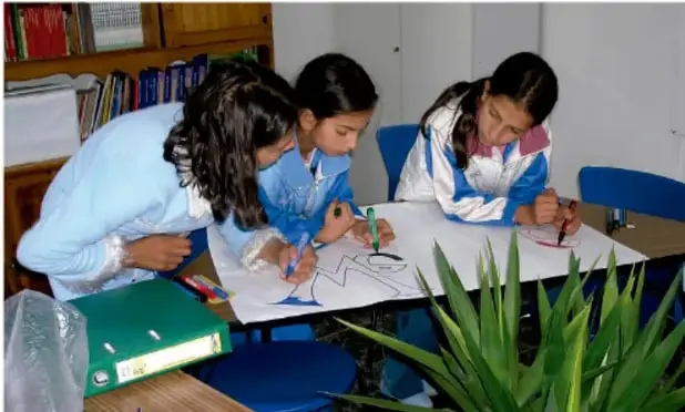 Fotografía de tres estudiantes con uniforme escolar trabajando de pie sobre una mesa; están dibujando y escribiendo con plumones en hojas blancas (láminas). Sobre la mesa hay un carpeta verde, materiales escolares y, al frente, una planta de hojas largas y verdes. El fondo muestra un estante con libros y revistas.