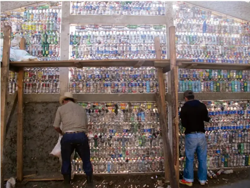 Fotografía de una construcción donde dos trabajadores colocan cientos de botellas de plástico de colores como módulos de una pared, sobre una estructura de madera; los llenan con material de relleno.
